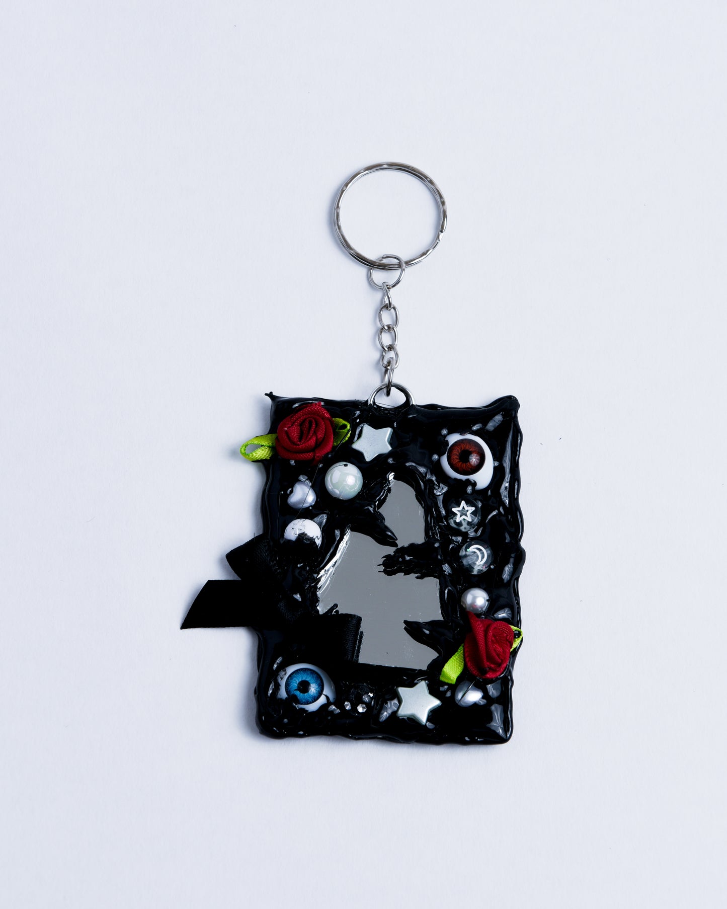 backside of black hot glue frame with beads, pearls, and small fabric bows surrounding the broken mirror piece on the back. Stainless steel rings serve as the keychain part, holding the piece together.