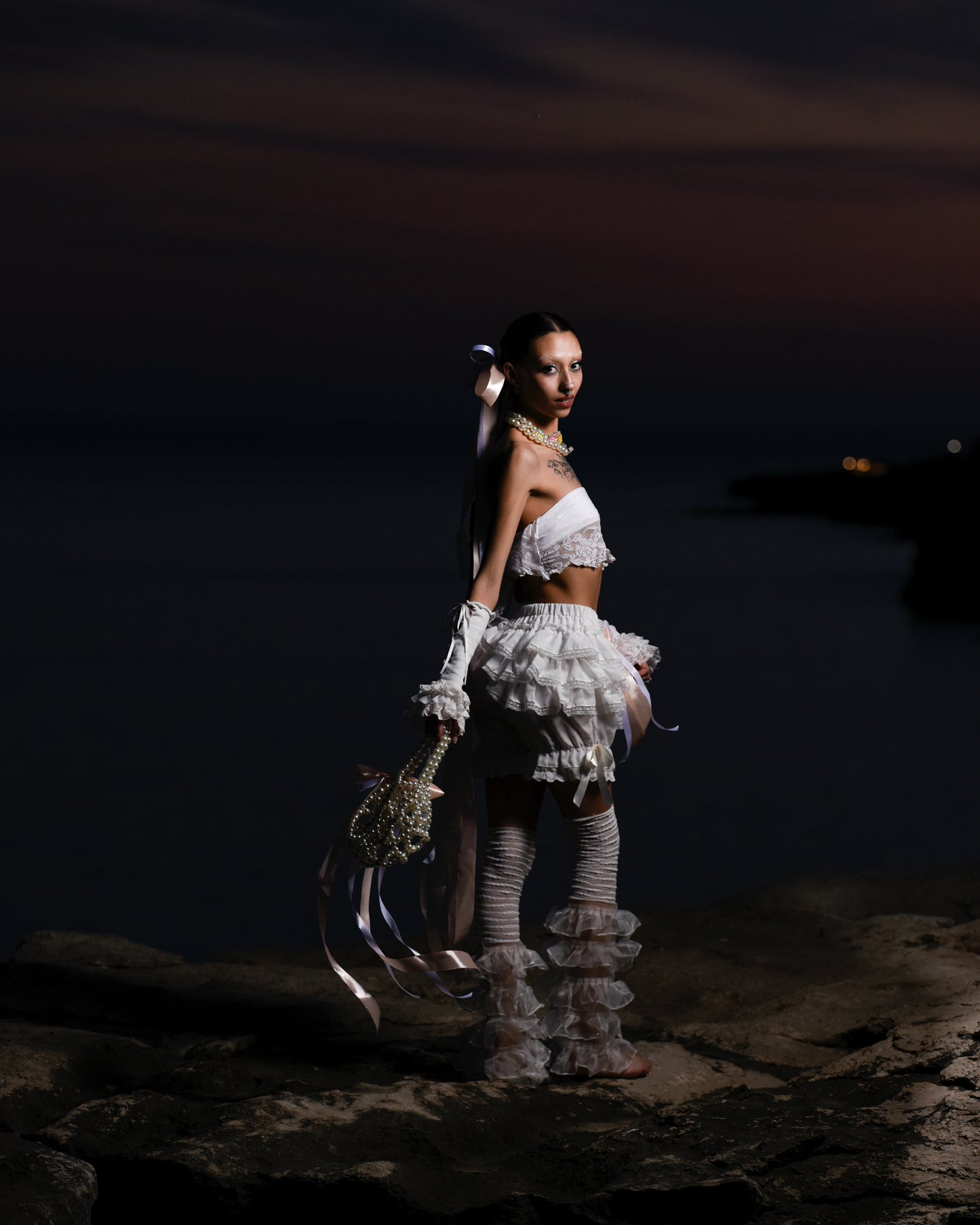 Model standing on a cliff in a white lace outfit, long bows floating from her head, with a pearl handbag swinging from her arm, featuring a small bow.