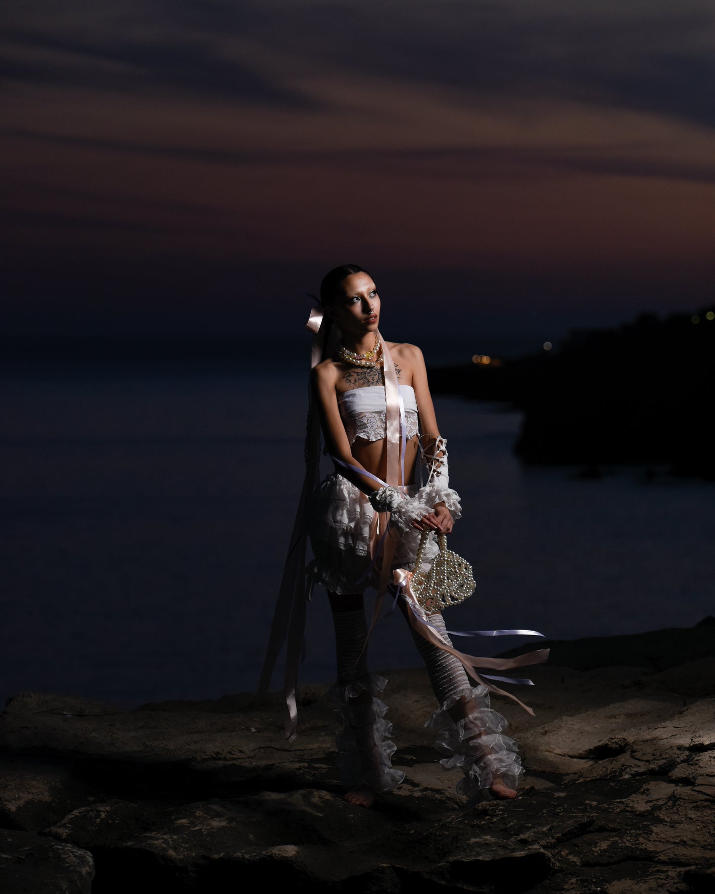 Model standing on a cliff in a white lace outfit, long bows floating from her head, with a pearl handbag swinging from her arm, featuring a small bow.