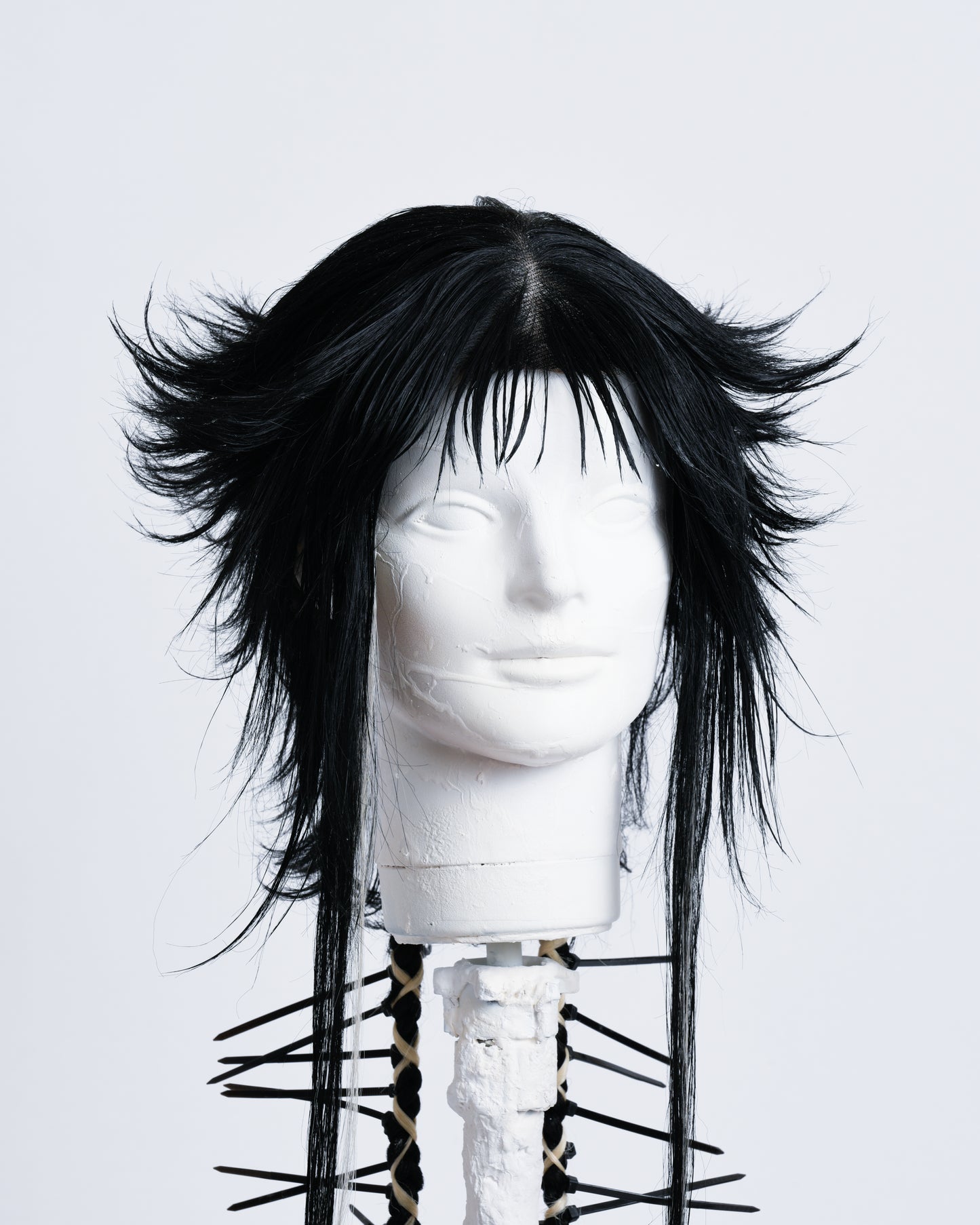 closeup of WIG Ren is a 70cm long anime cut black wig with bangs and two braids in the back with white highlights braided in and black cable ties coming out .