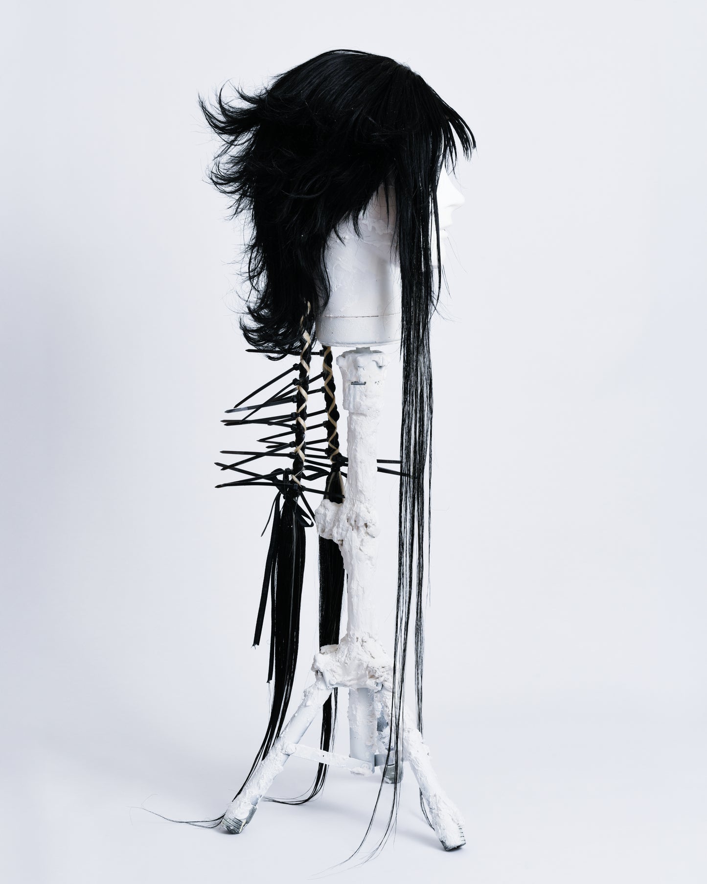 side profile of WIG Ren is a 70cm long anime cut black wig with bangs and two braids in the back with white highlights braided in and black cable ties coming out .